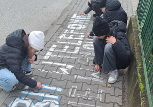 Dzieci piszą na chodniku hasło kredą.