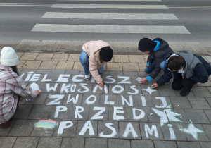 Dzieci piszą kredą hasło na chodniku przy szkole.