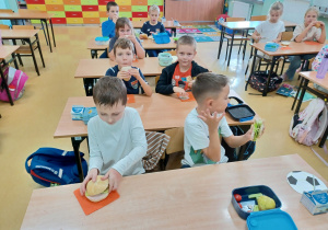 Pierwszoklasiści siedzą w ławkach i jedzą śniadanie.