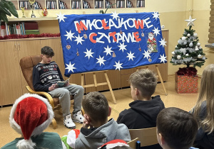 Uczniowie słuchają fragmentów książki czytanych przez ucznia klasy ósmej.