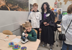 Przebrani siódmoklasiści stoją za chłopcem, który patrzy przez mikroskop.