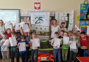 Dzieci z grupy "0" stoją z Paniami prowadzącymi zajęcia przy tablicy i trzymają prace wykonane podczas tych zajęć.