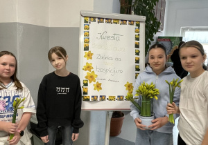 Dziewczynki stoją z żonkilami przy tablicy informującej o kweście.