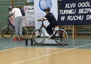 Jeden z chłopców jedzie po torze przeszkód, drugi pochyla się przy rowerze.