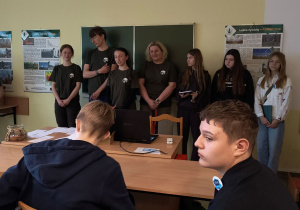 Pod tablicą stoją organizatorzy konkursu oraz nagrodzone uczennice, a uczniowie siedzą w ławkach.