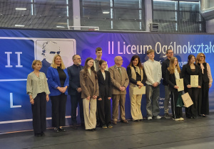Szymon Rudzki stoi pomiędzy uczniami wyróżnionymi w konkursie oraz ich opiekunami i organizatorami.