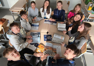 Uczniowie siedzą w kole przy ławkach i przeglądają prospekty.
