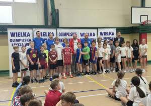 Uczniowie klas czwartych stoją z Olimpijczykami i organizatorami imprezy.