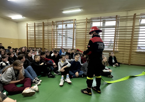 Młody strażak prezentuje uczniom wąż do gaszenia pożarów