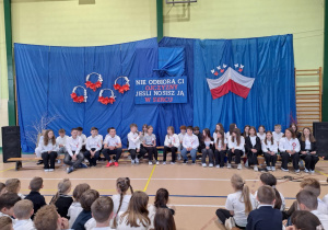 Uczniowie klasy VIa oraz członkowie zespołu wokalnego na tle odświętnej dekoracji w hali sportowej